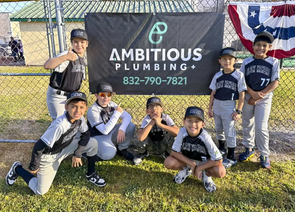 Community involvement in League City youth sponsorship. Local Little League baseball team posing in front of an Ambitious Plumbing sponsorship banner on a sports field fence