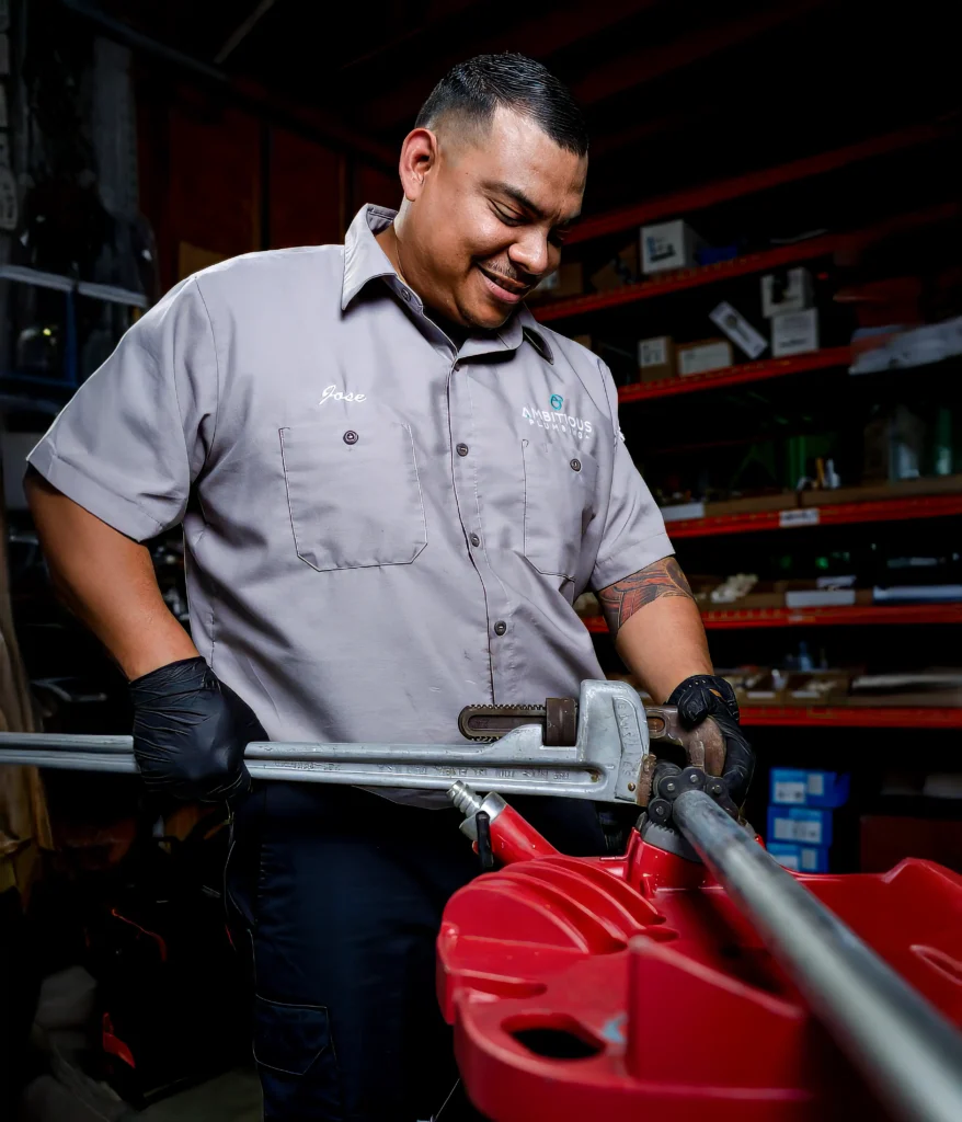 Ambitious Plumbing team member Jose using a heavy-duty pipe wrench to prep a metal pipe in the workshop, serving League City, Bellaire, and Greater Houston. Plumbing Services in League City by Ambitious Plumbing.